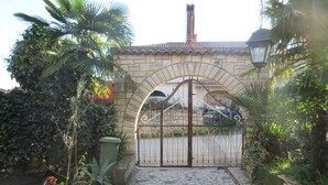 Un chemin de pierre mène à une entrée fermée par un portail, surmontée d’un toit en tuiles, et est entouré d’une végétation luxuriante et de plantes en pot.