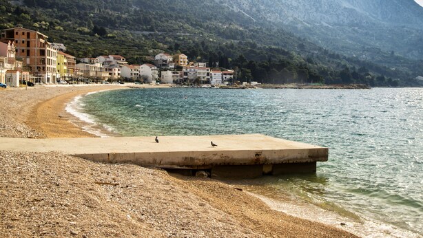 A coastal area with a pebbled beach, a concrete pier, and buildings in the background. - House Lea (Igrane)
