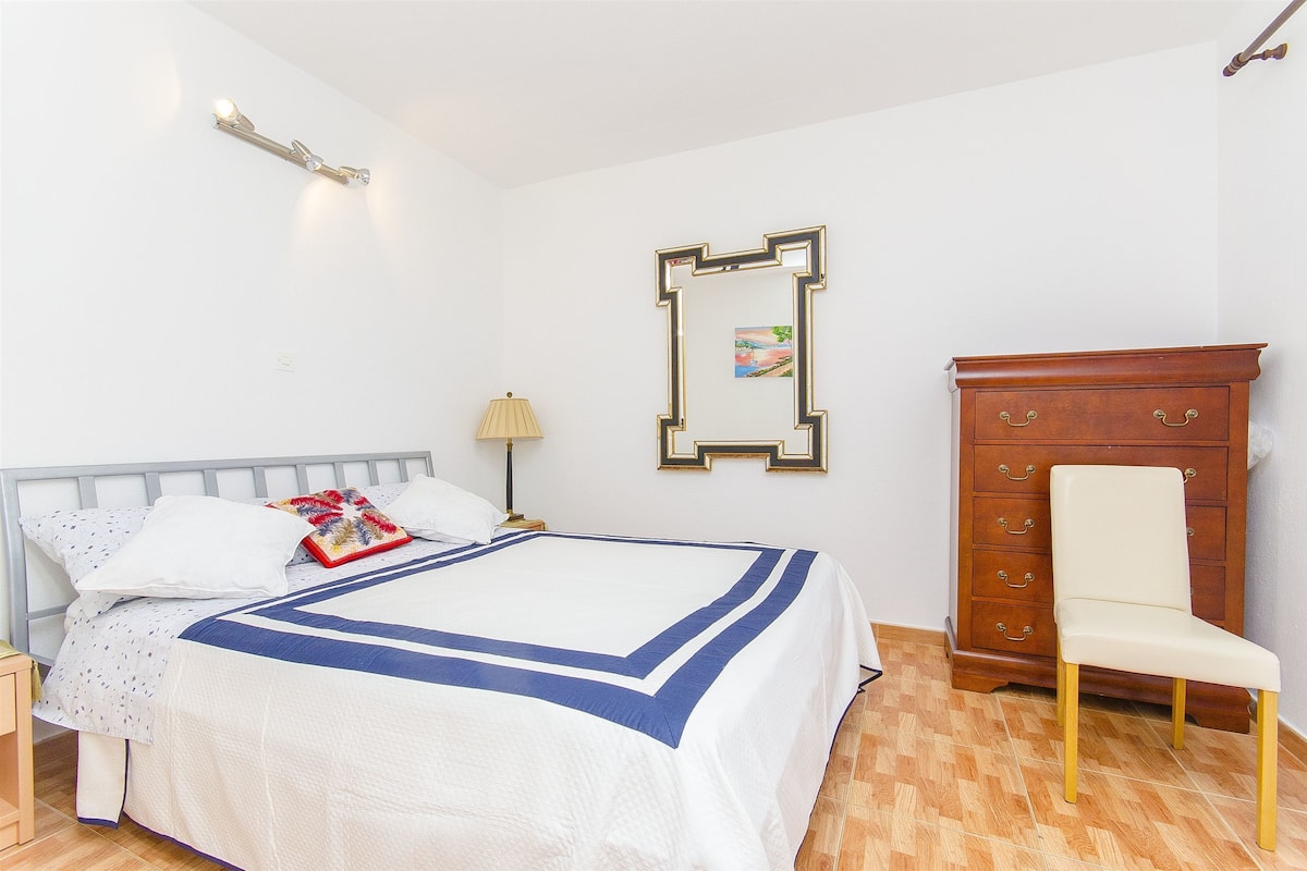 Una cama bien hecha con una colcha azul y blanca, una mesita de noche con una lámpara, un aparador de madera con cajones y una silla.