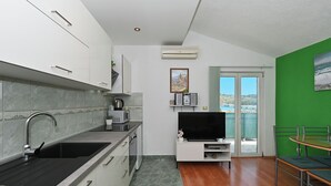 A modern kitchen with a built-in stove, microwave, and sink. There is a flat-screen TV on a white stand, and a dining area with a green wall and a view of the sea. - Apartments Marija (Trogir)
