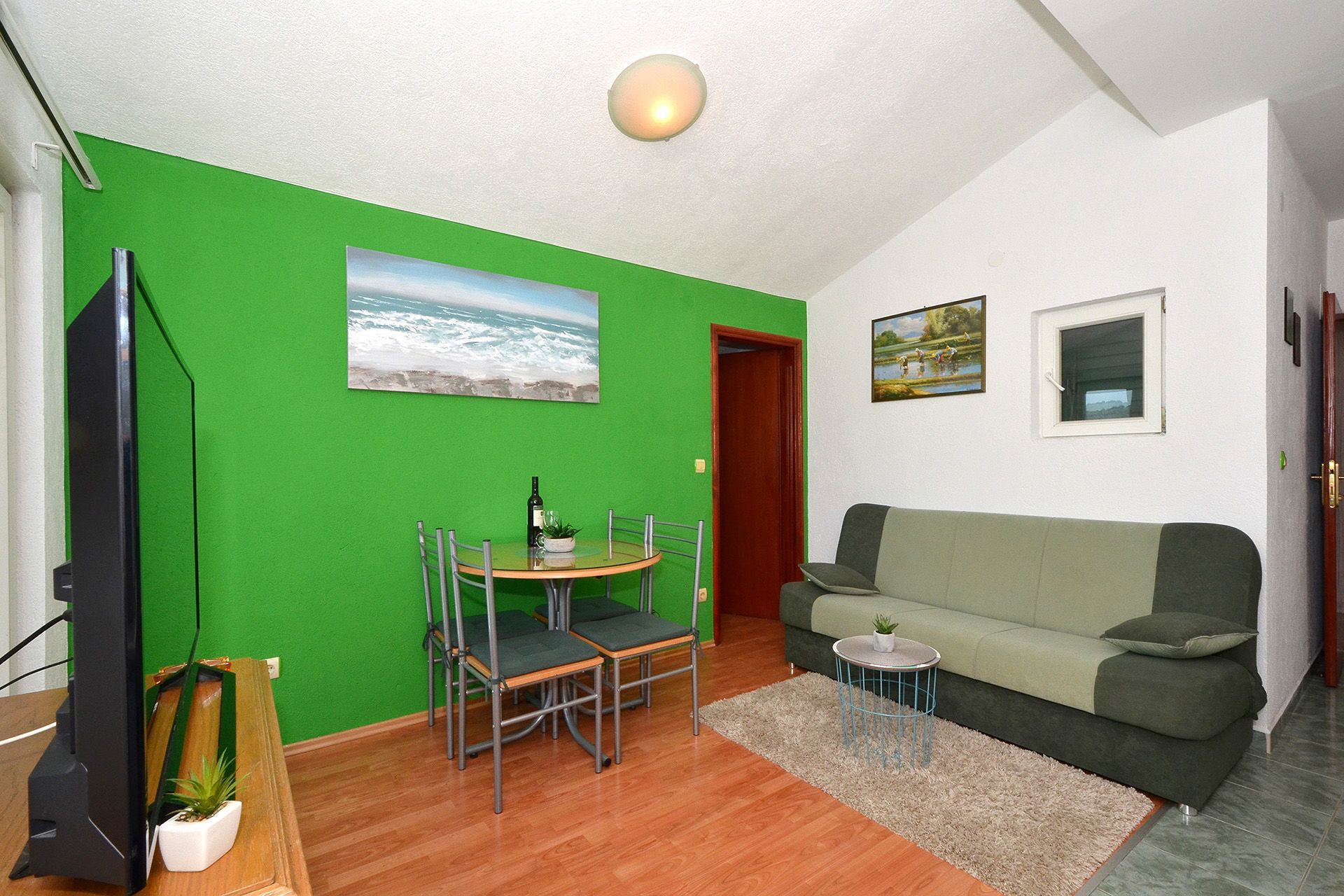 A living room with a green wall, a wooden floor, a green sofa, a small round table, and a TV.