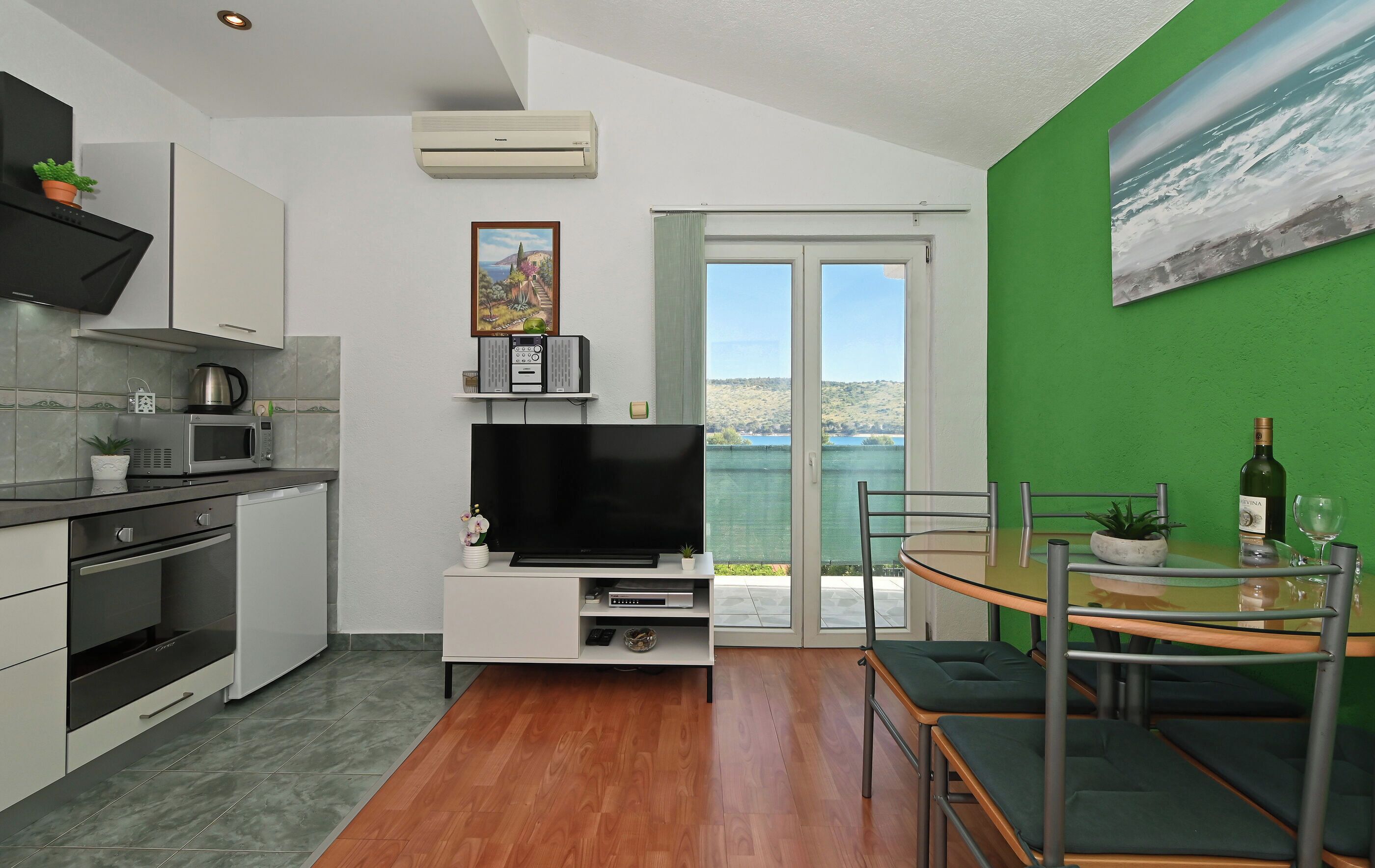 Cocina moderna con televisor de pantalla plana, comedor con mesa y sillas, pared de acento verde y vista a un cuerpo de agua a través de una puerta de vidrio.