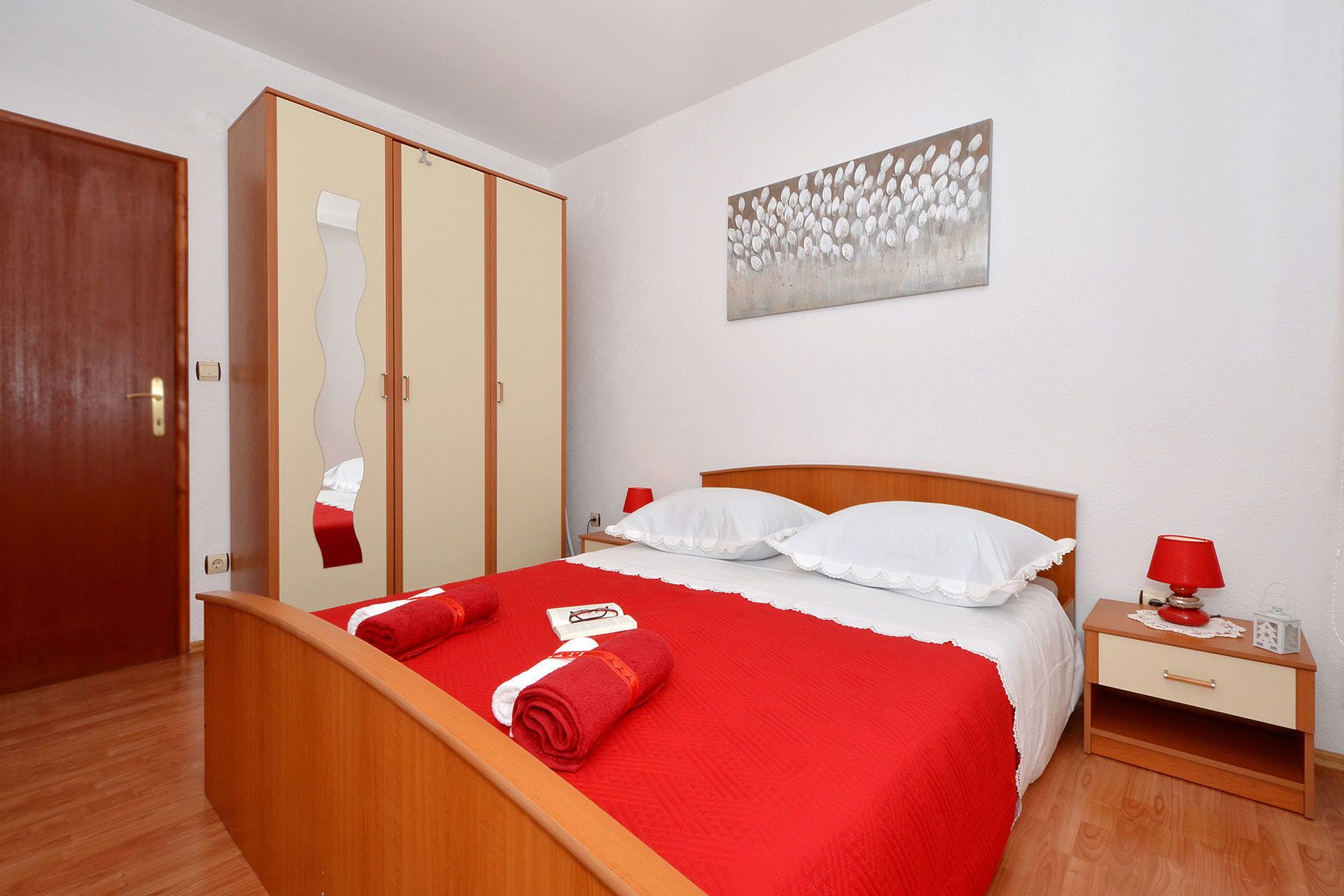 A hotel room with a wooden bed, red bedding, a nightstand with a red lamp, and a wardrobe with a mirror.
