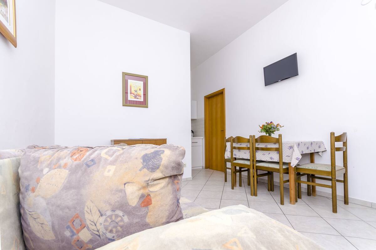 A living room with a sofa, a dining table with chairs, a TV mounted on the wall, and a door leading to another room.