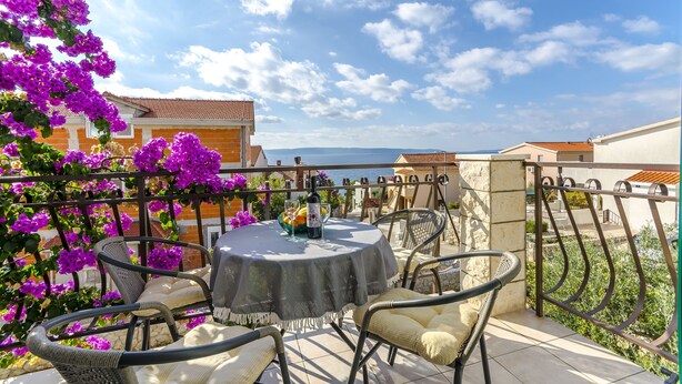 A terrace with a round table and chairs, surrounded by purple flowers, overlooking a coastal area. - Apartments Mate (Okrug Gornji)