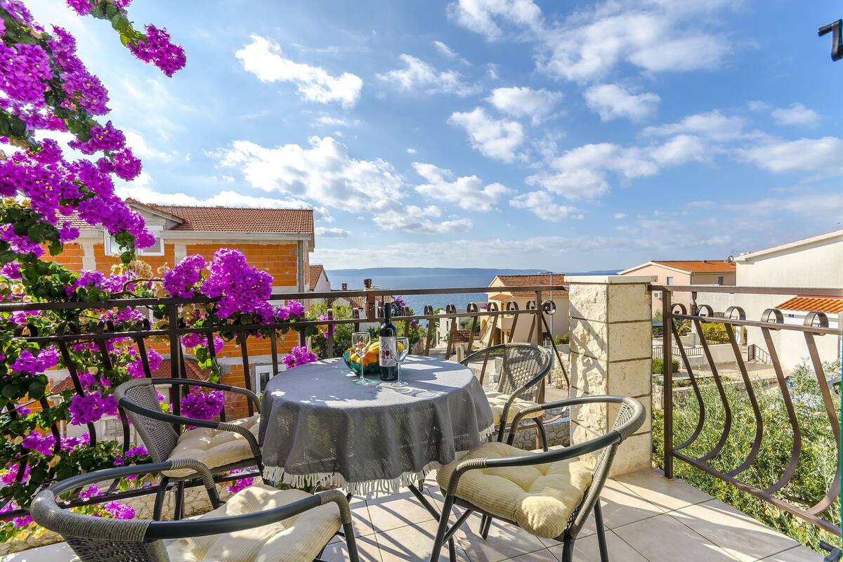 A terrace with a round table and chairs, surrounded by purple flowers, overlooking a coastal area.