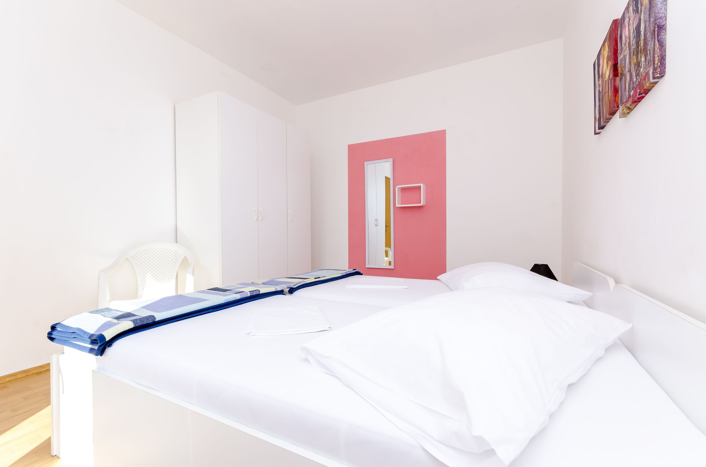 Uma cama de solteiro com roupa de cama branca, uma cadeira branca e uma parede cor-de-rosa com espelho e obras de arte.
