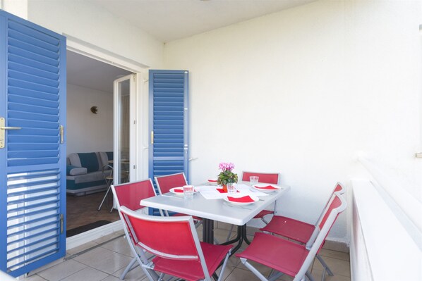 Un coin repas avec une table blanche dressée pour quatre personnes, des chaises rouges et des volets bleus aux murs.