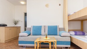 Ένας μικρός και λειτουργικός χώρος διαβίωσης με έναν καναπέ, ένα μικρό τραπέζι, ένα κρεβάτι-κουκέτα και μια μικρή κουζίνα.