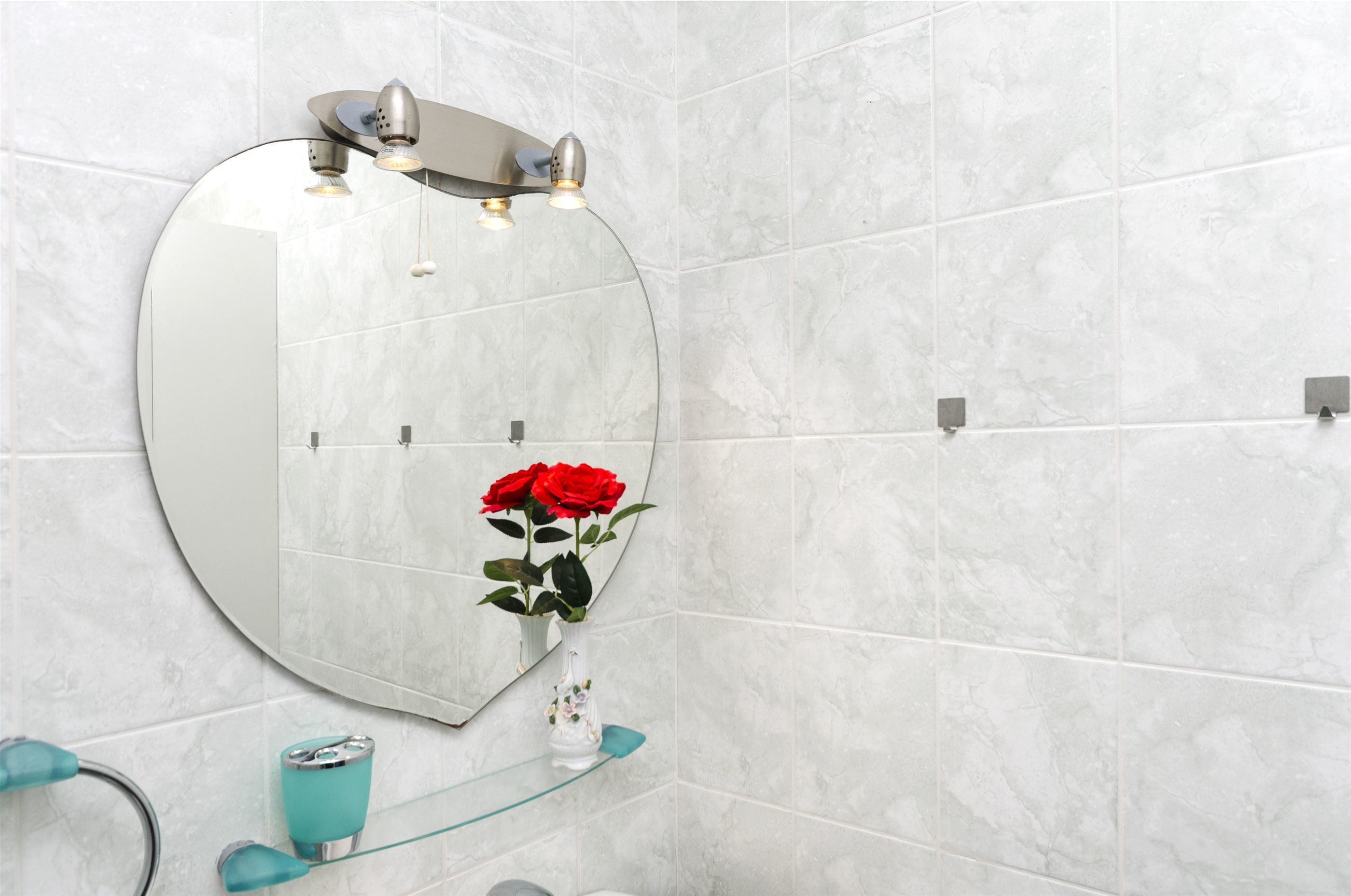 A bathroom with a heart-shaped mirror, a vase with red roses, and a glass shelf.