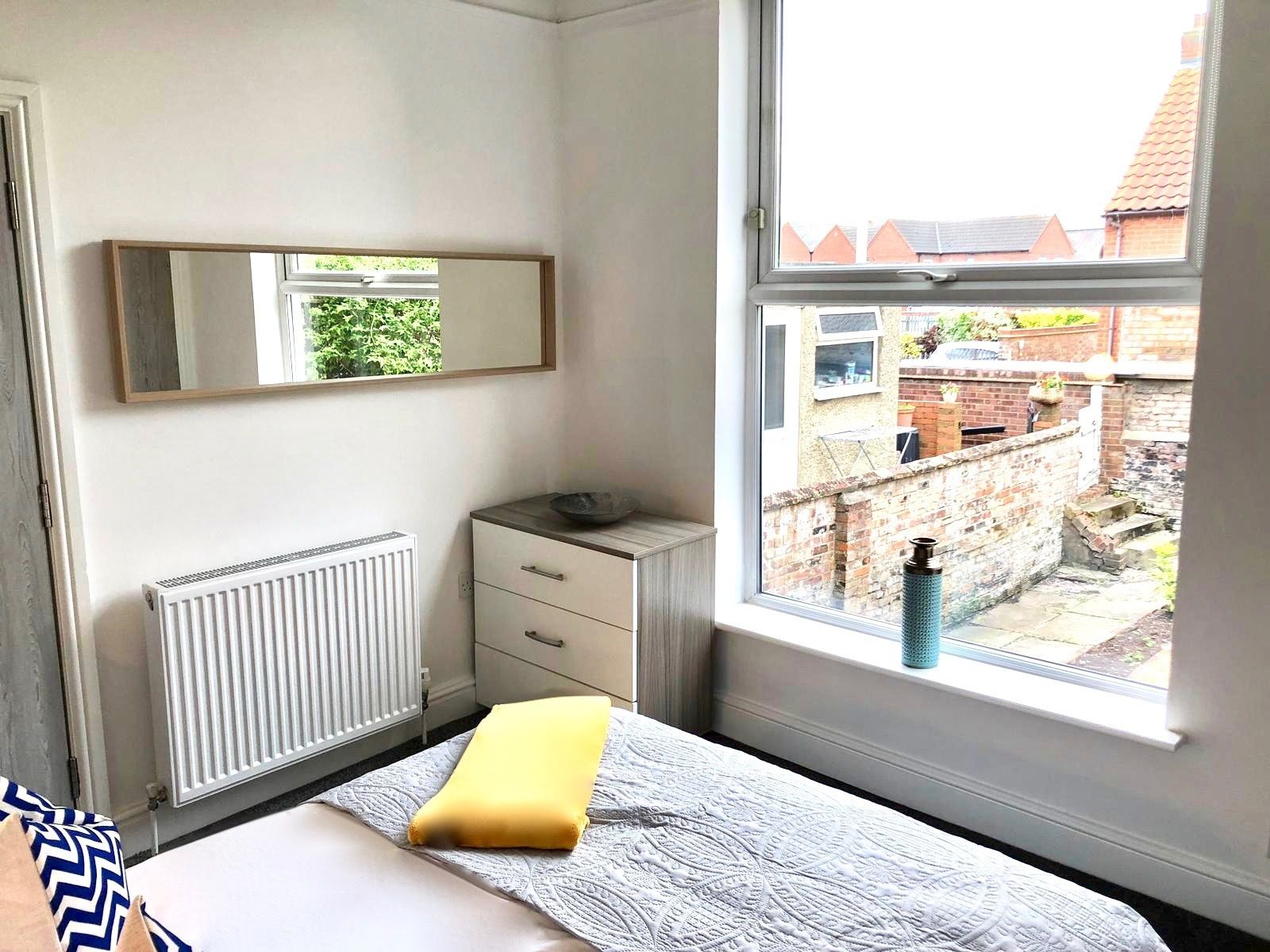 Un dormitorio con cama, una mesita de noche con un cuenco, un radiador, y una ventana que ofrece vistas a un muro de ladrillo y vegetación.