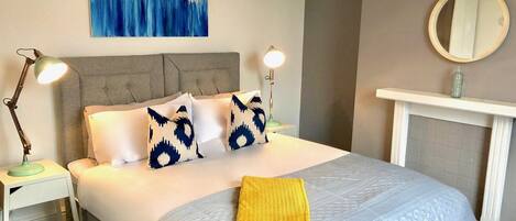 Uma cama bem arrumada com almofadas estampadas, duas luminárias de cabeceira, uma obra de arte na parede e uma lareira com um espelho acima.