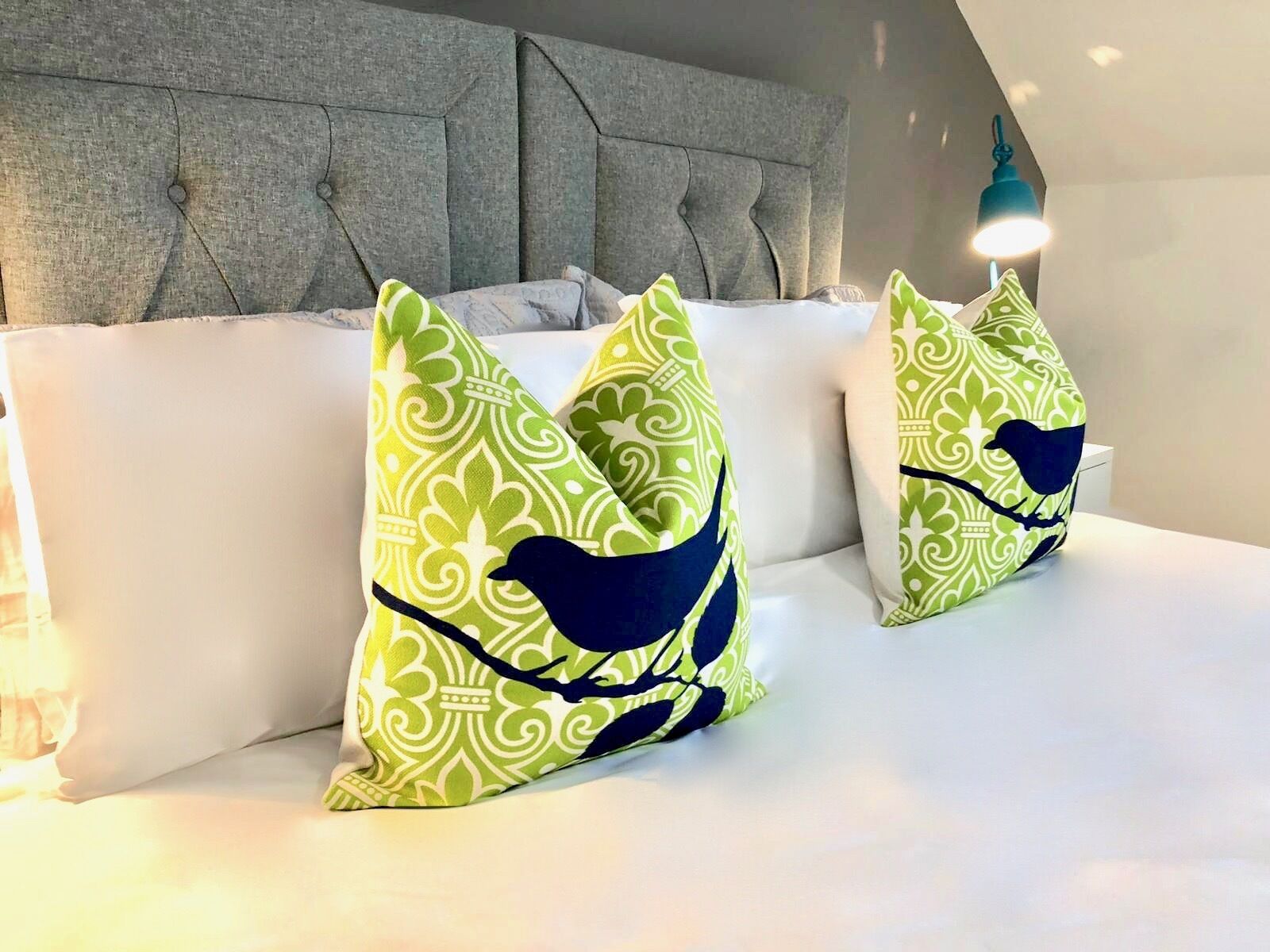 Una cama con cabecera tapizada de gris, dos almohadas verdes estampadas con diseño de pájaros y un colchón blanco.