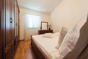 A bedroom with a wooden wardrobe, a bed with white bedding, a wooden dresser, a mirror, and a window with sheer curtains.