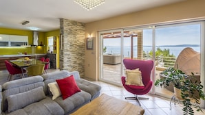 A modern living room with a grey sofa, red and white cushions, a wooden coffee table, and a red armchair. A glass dining table with chairs is in the background. A large sliding glass door leads to a balcony with a view of the sea. - Apartment Paradise View (Okrug Gornji)