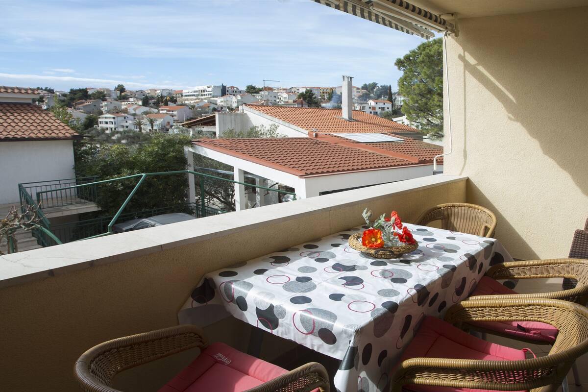 A balcony with a table and wicker chairs, overlooking a residential area with red-tiled roofs.