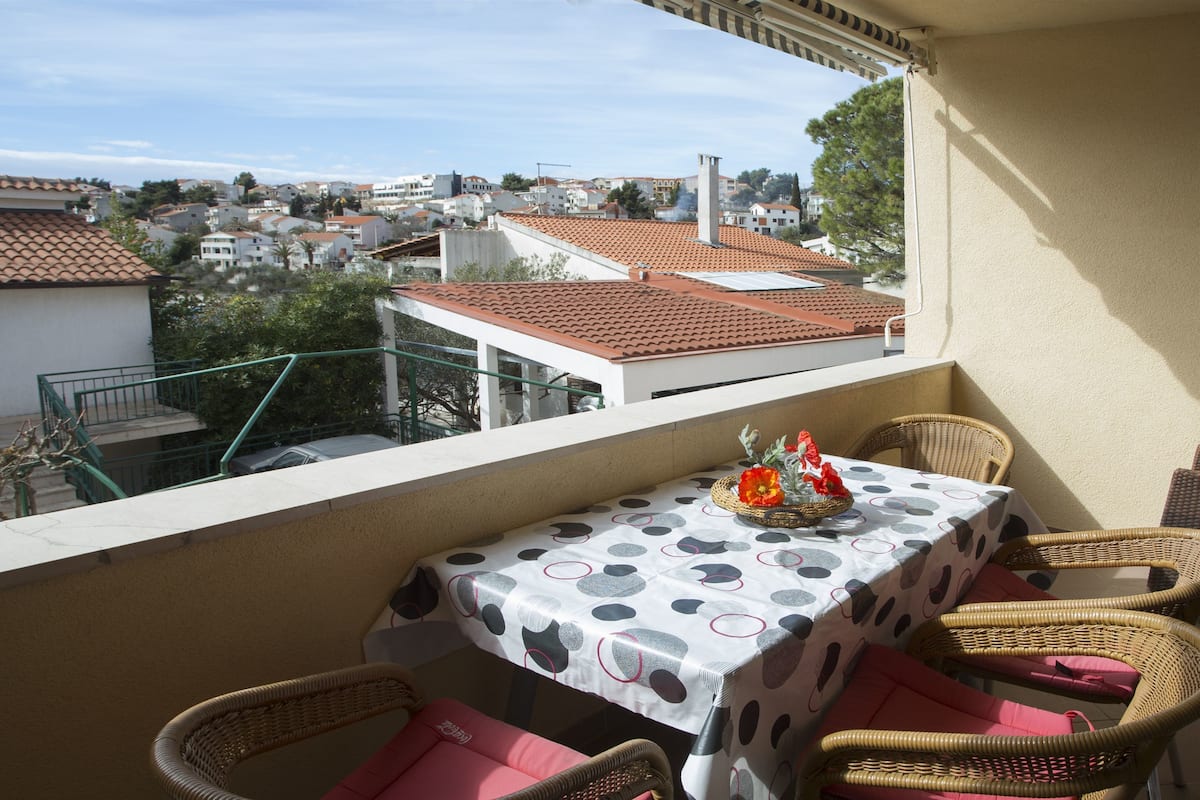 Un balcón con una mesa y sillas de mimbre, con vistas a una zona residencial de tejados rojos.
