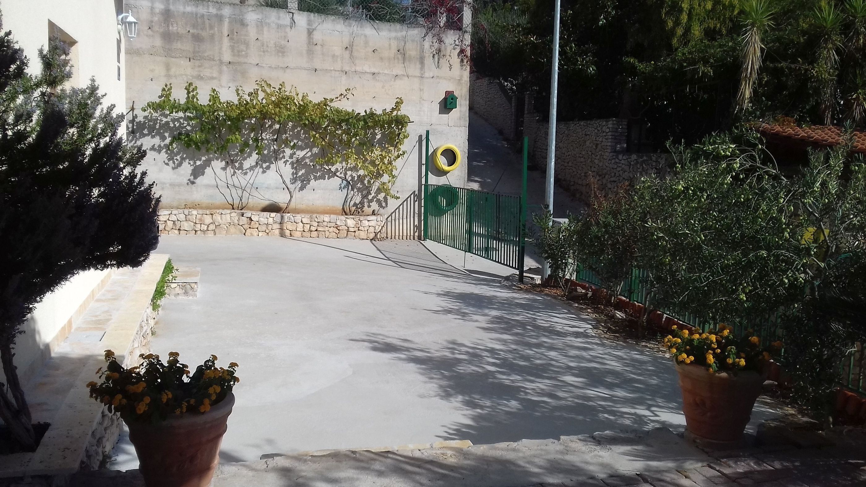 Un vialetto lastricato conduce a un edificio con un muro in pietra e una recinzione verde. Ai lati del percorso si trovano vasi con fiori gialli.