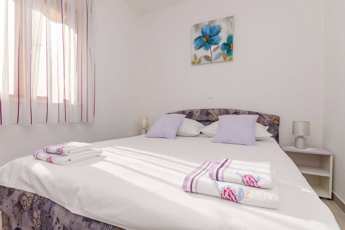 Un lit bien fait, avec du linge de lit blanc et des serviettes pliées, une tête de lit ornée d’un motif floral et un tableau représentant des fleurs bleues accroché au mur.