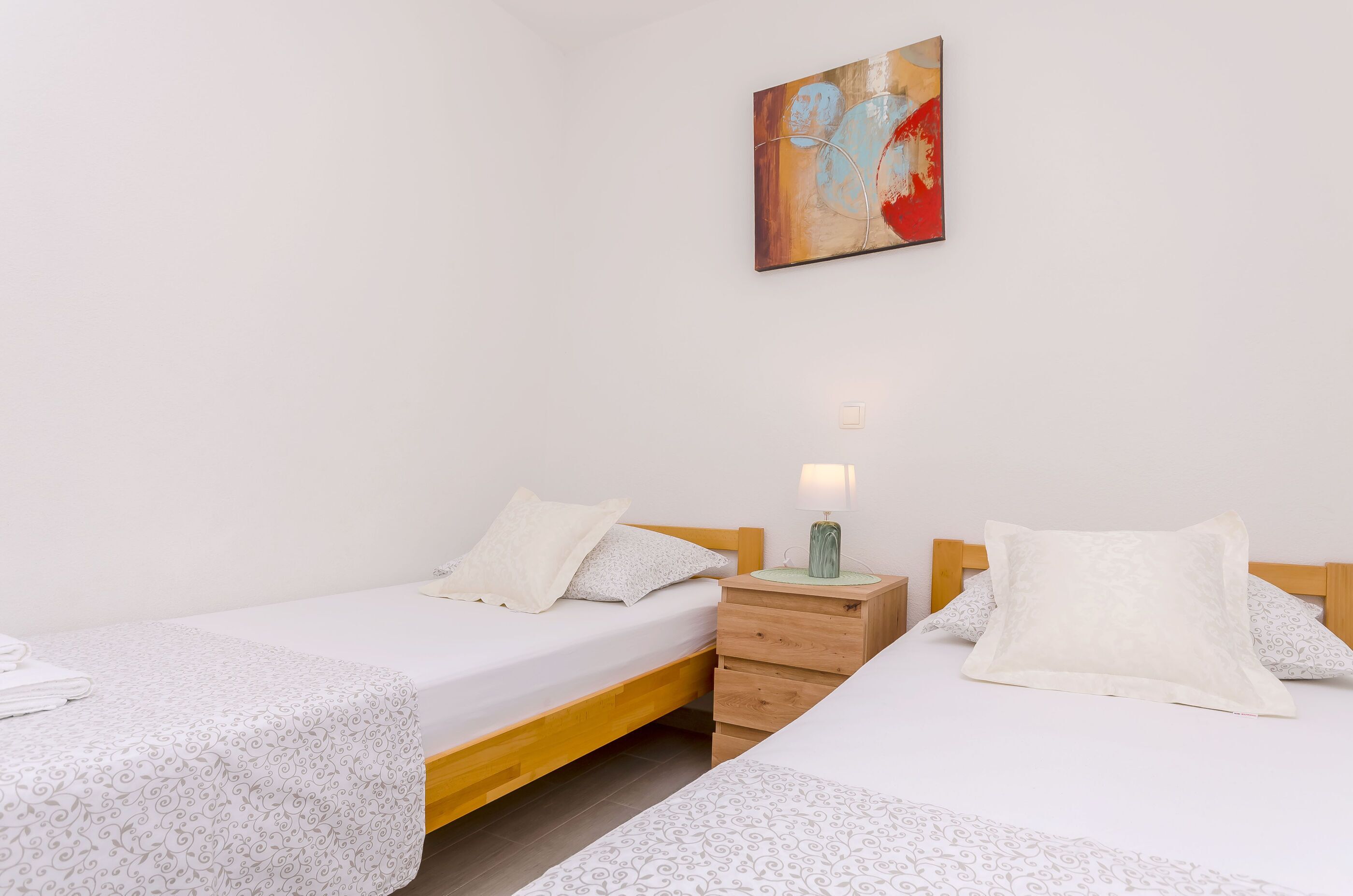 A bedroom with two single beds, a wooden nightstand with a lamp, and a painting on the wall.