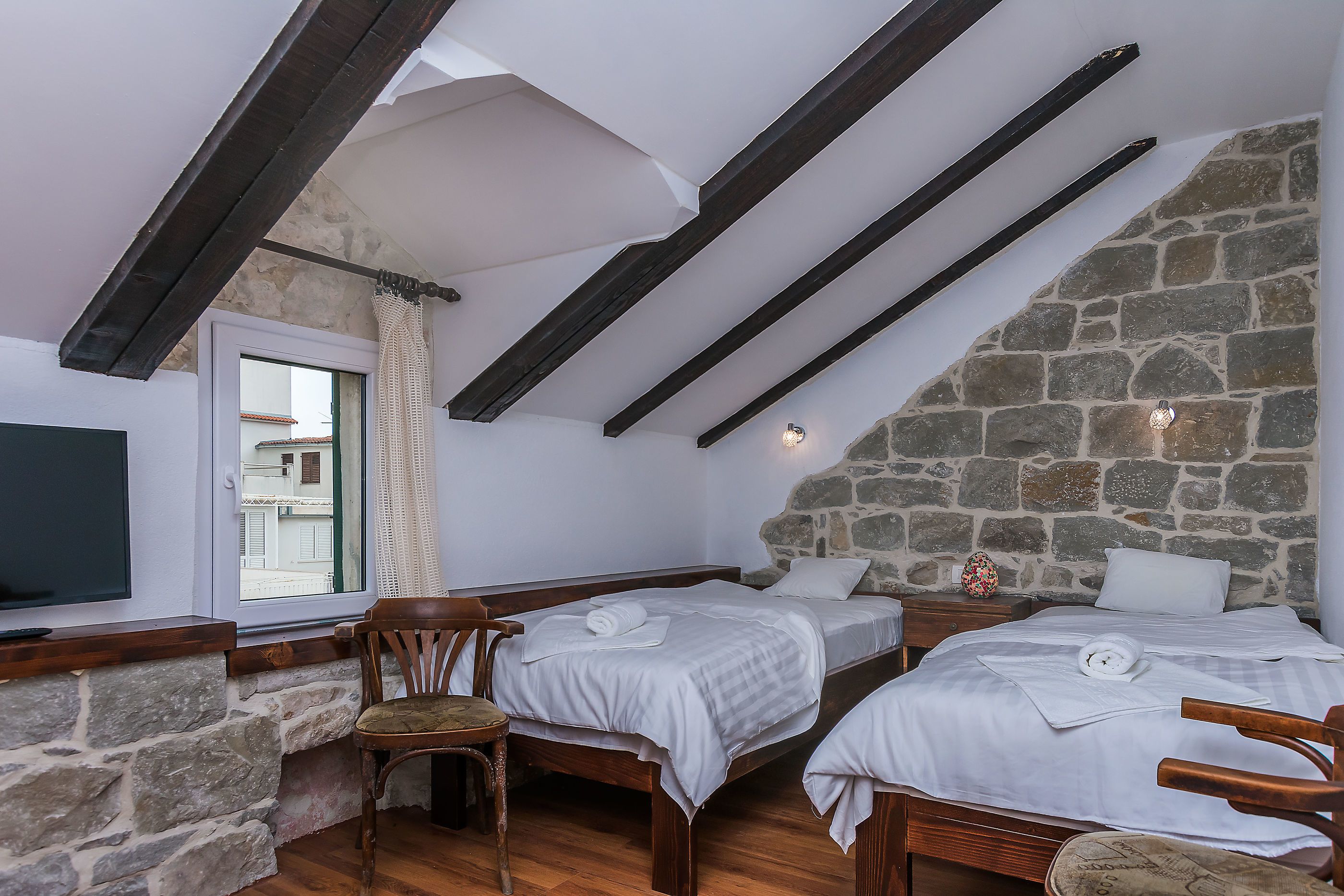 A room with two beds, wooden beams, stone walls, and a window with a view.