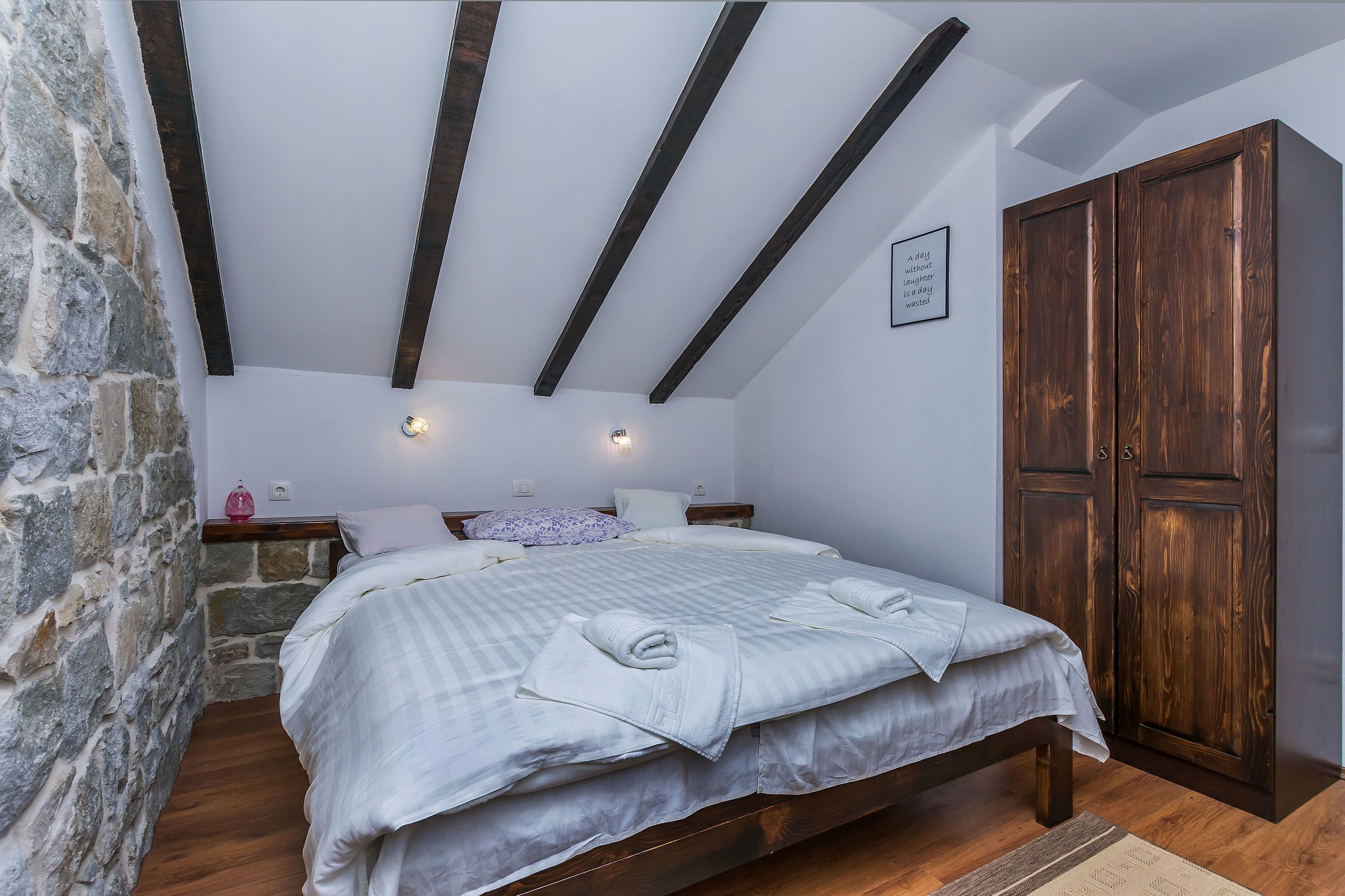 A bedroom with a stone wall, a wooden bed, a wardrobe, and a sloped ceiling with wooden beams.