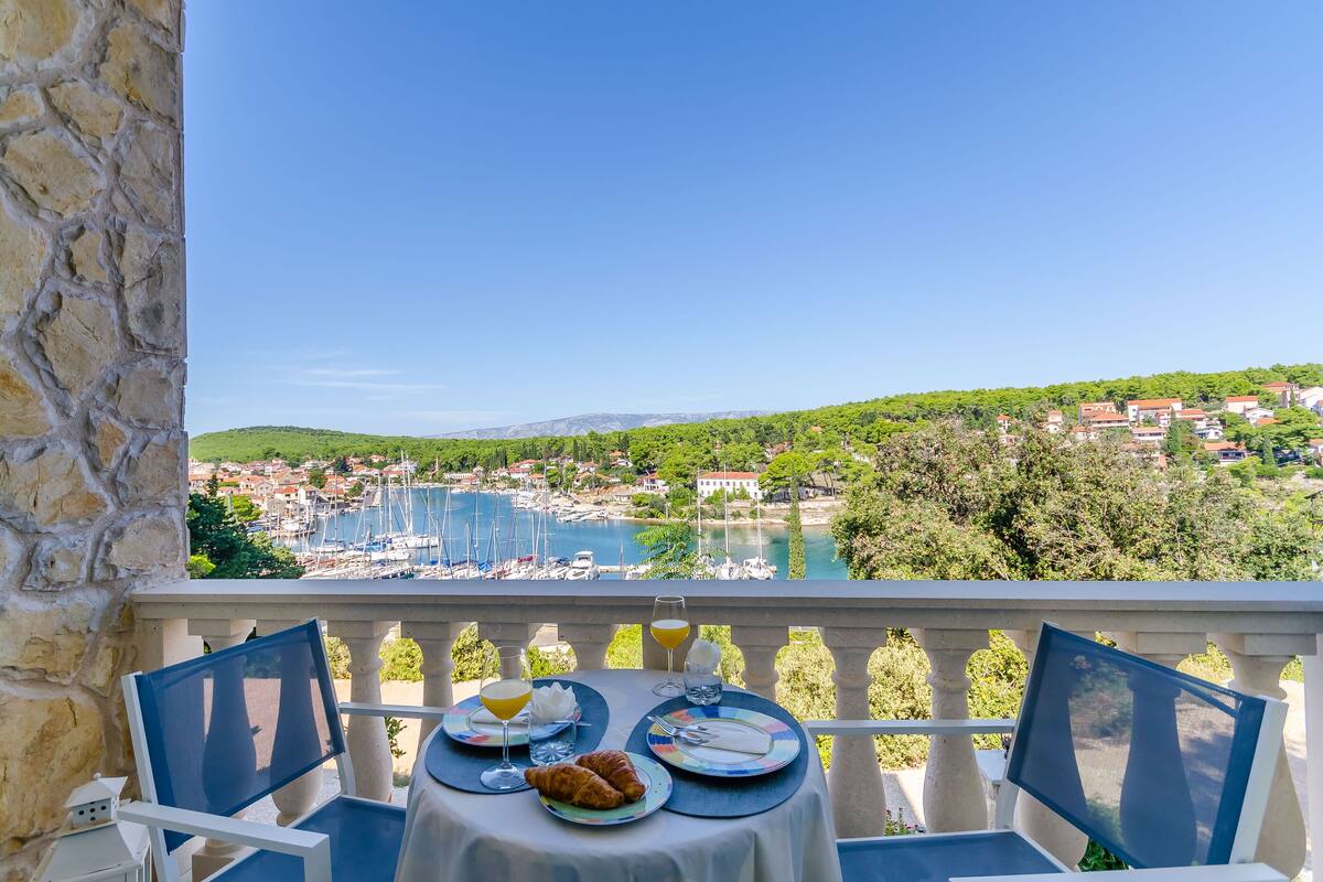 A balcony with a table set for breakfast overlooking a marina and a coastal town.