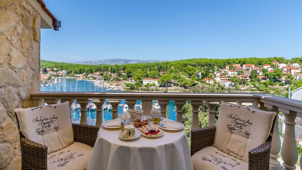 A balcony with wicker chairs and a table set for breakfast, overlooking a marina and a coastal town. - Apartments Vila Jelka (Vrboska)