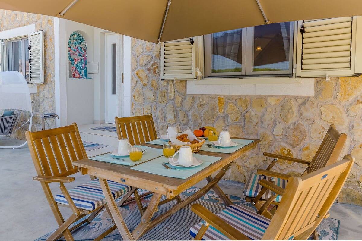 A stone-walled room with a wooden table set for breakfast, including fruits and a basket of bread.