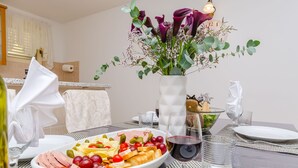 A dining table set with a variety of food, including a bowl of fruit and cheese, and a glass of red wine. There is a vase with flowers in the background. - Apartments Vila Jelka (Vrboska)