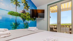 A hotel room with a large bed, a flat-screen TV mounted on the wall, and a view of a pool and outdoor seating area. - Apartments Vila Jelka (Vrboska)