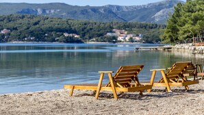 A beach with wooden lounge chairs and a calm lake, surrounded by mountains and houses in the background. - Apartments Vila Jelka (Vrboska)