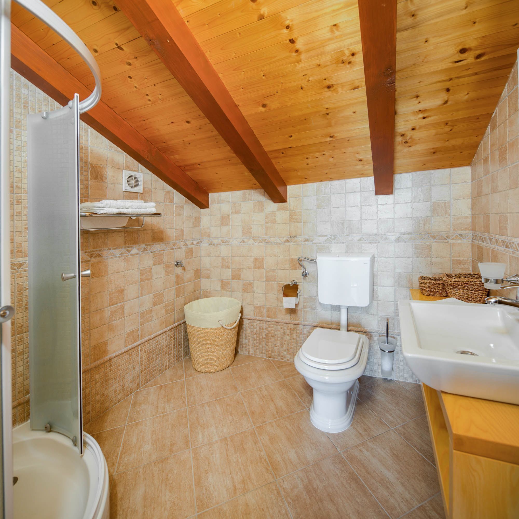 A bathroom with a shower, toilet, sink, and a tiled wall with a wooden ceiling.