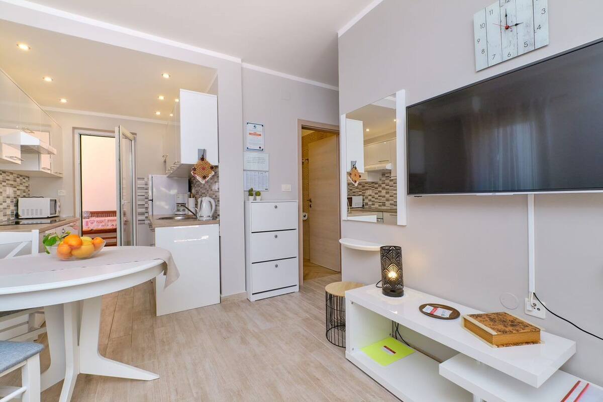 Une cuisine moderne avec des meubles blancs, une table à manger ornée de fruits et un téléviseur fixé au mur.