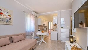 Una sala de estar moderna con un sofá, una mesa de comedor con sillas, una cocina al fondo y un televisor montado en la pared.