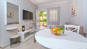 Una sala de estar moderna con un televisor, una mesa de comedor blanca con frutero, un sofá blanco y una mesita de noche con lámpara.