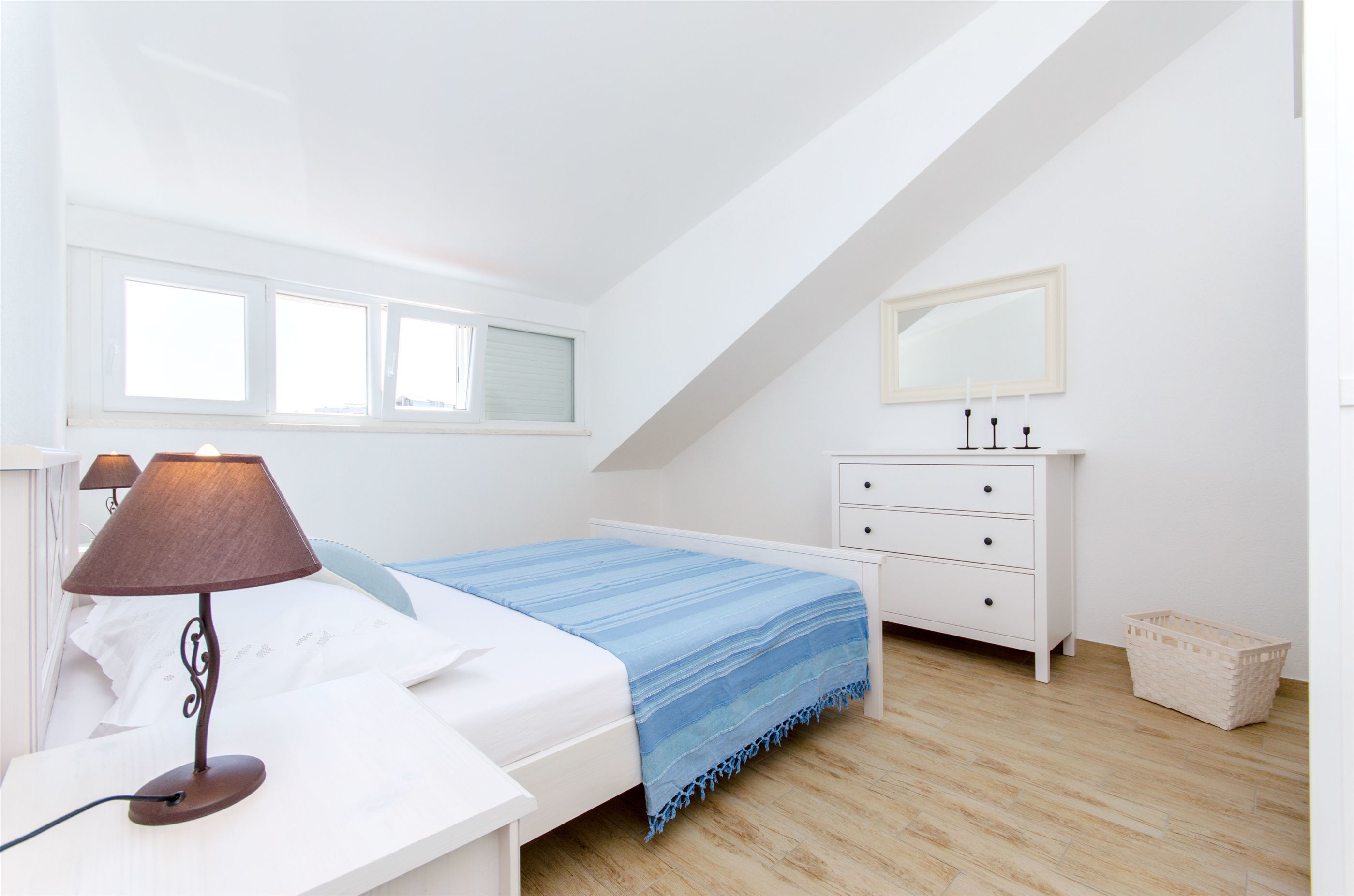 A bedroom with a bed, a bedside table with a lamp, a dresser, a mirror, and a window.