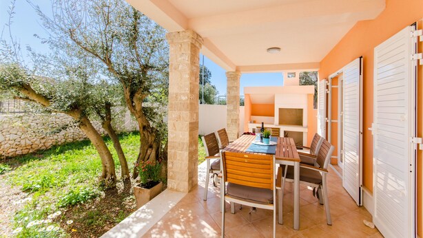 Un patio esterno coperto con tavolo e sedie, colonne in pietra e vista su un giardino con alberi e un muro in pietra.