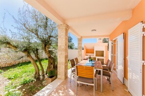 A covered outdoor patio with a table and chairs, stone pillars, and a view of a garden with trees and a stone wall. - House Nikolina (Sevid)
