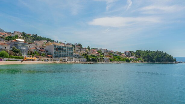 A coastal town with buildings along the waterfront, clear blue water, and a hilly backdrop. - Apartment Heaven (Strozanac)