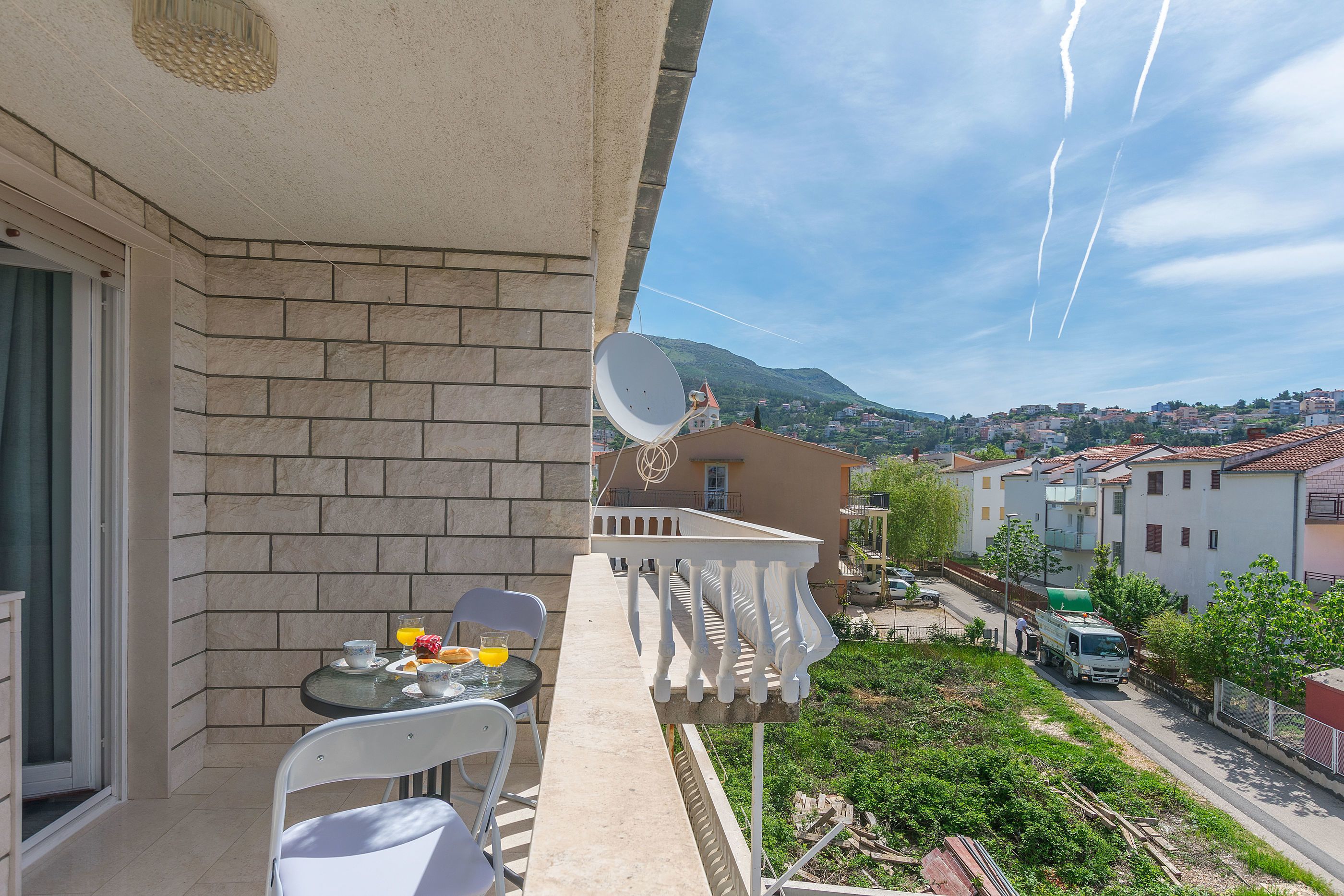 A balcony with a table and chairs, a satellite dish, and a view of a residential area with houses and a mountain in the background.