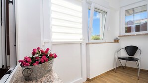 Une pièce avec un plancher en bois, une porte blanche, une fenêtre avec des stores, une chaise et un seau rempli de fleurs roses posé sur un comptoir en granit.