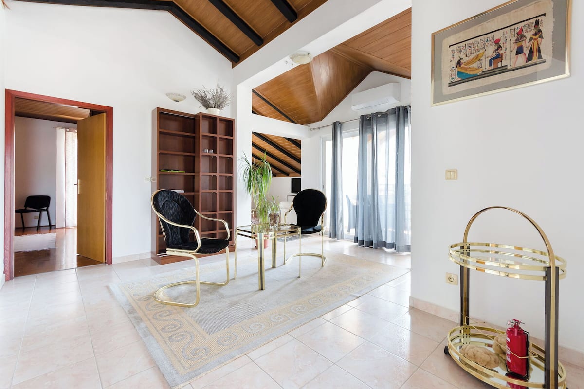 A spacious living room with a high ceiling, wooden beams, a large bookshelf, two black chairs, a small table with a plant, and a gold-framed side table with a red object.