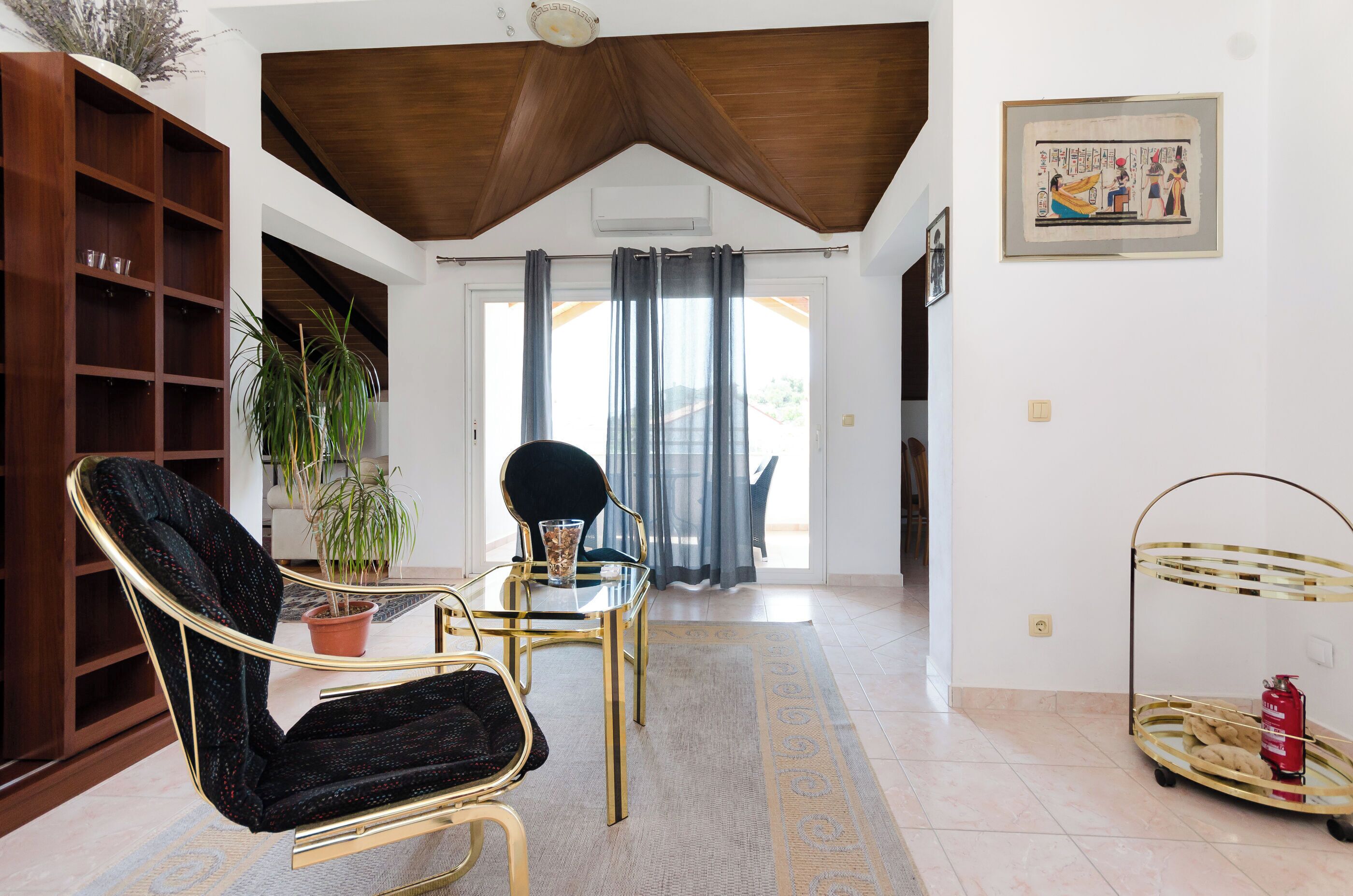 A modern living room with a wooden ceiling, a glass table, a black chair, a potted plant, and a fire extinguisher on a cart.