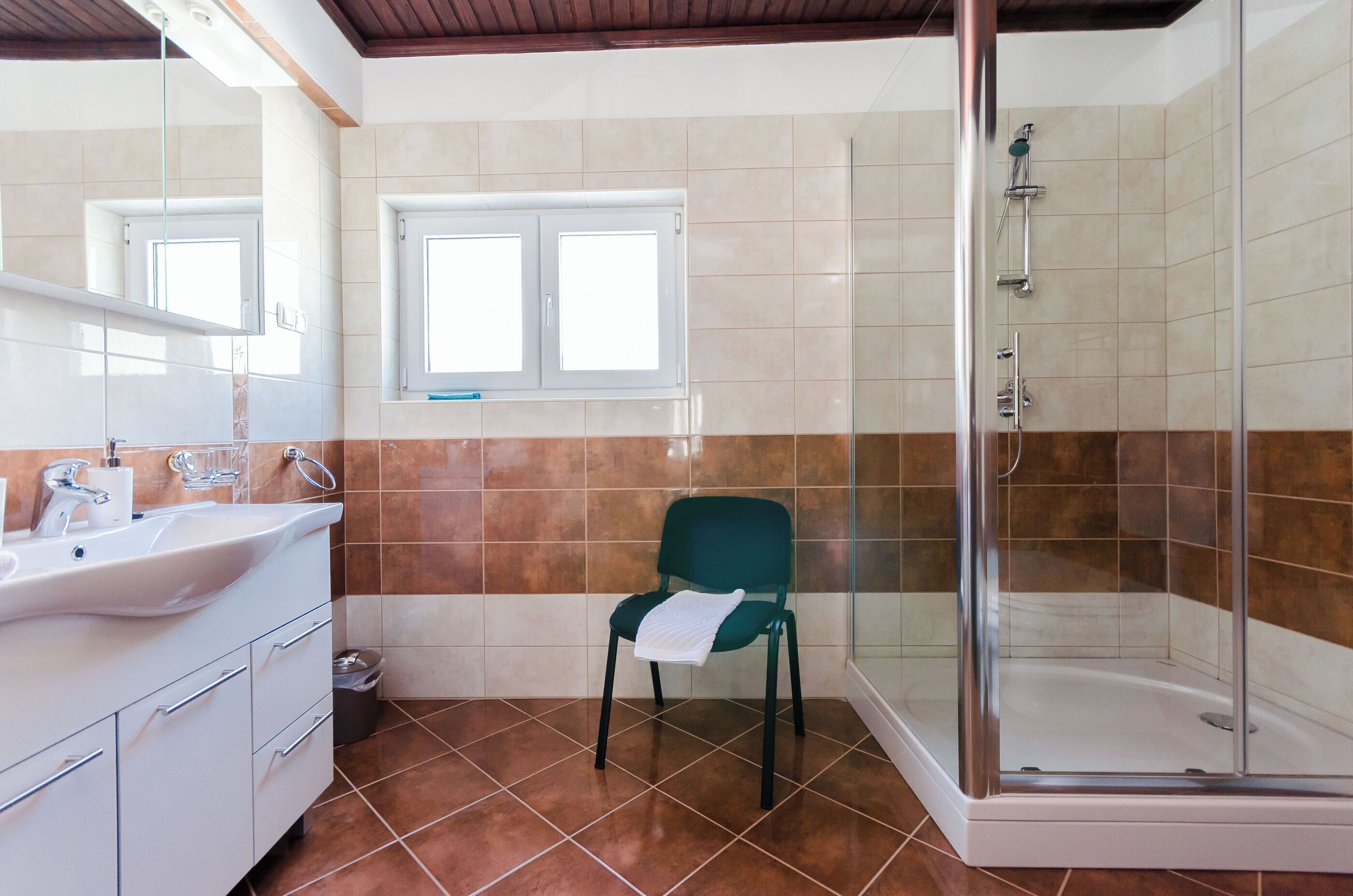 A bathroom with a shower, a sink, a chair, and a window.