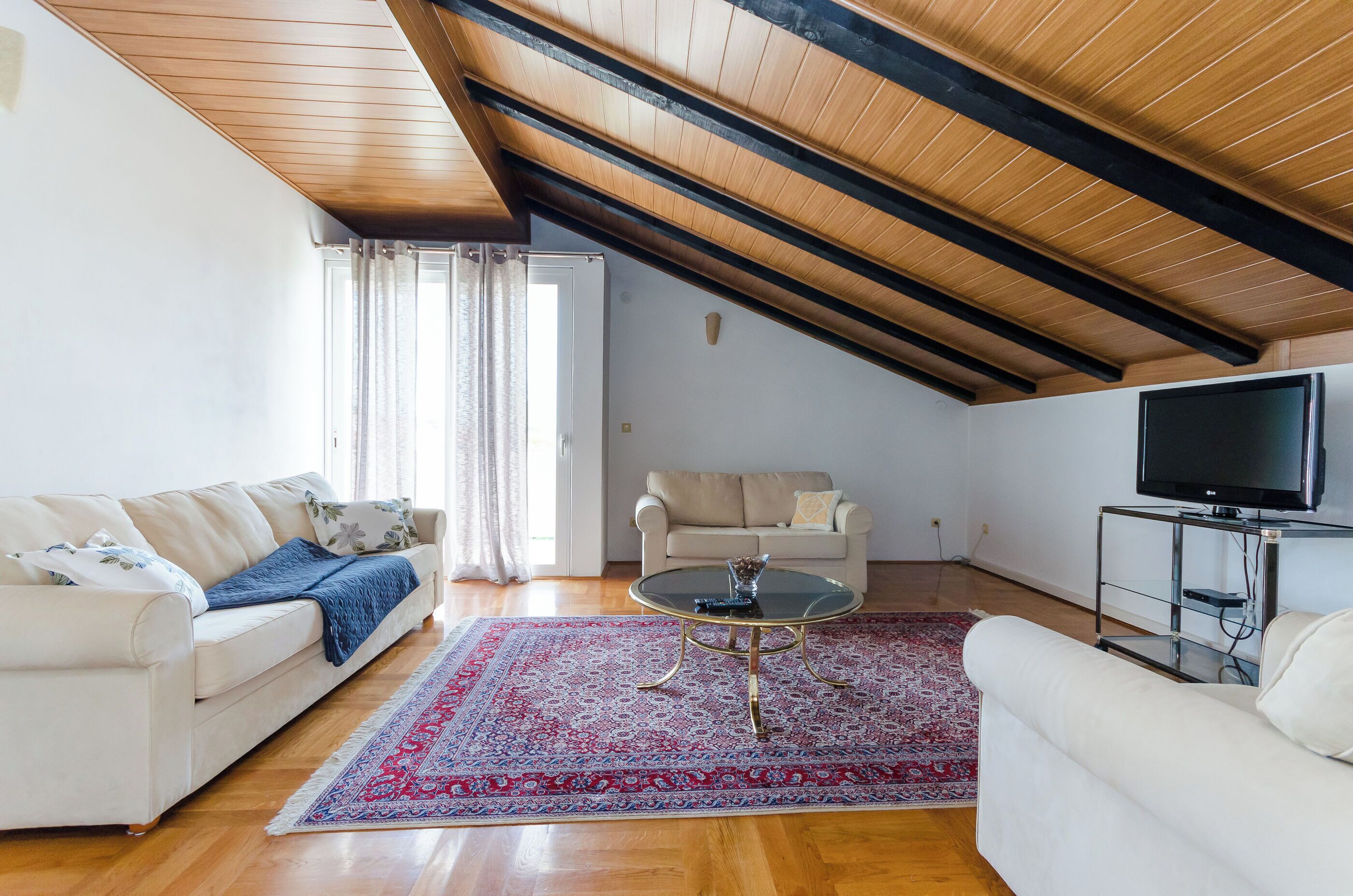 A living room with two white sofas, a glass coffee table, a red patterned rug, a television on a stand, and wooden beams on the ceiling.