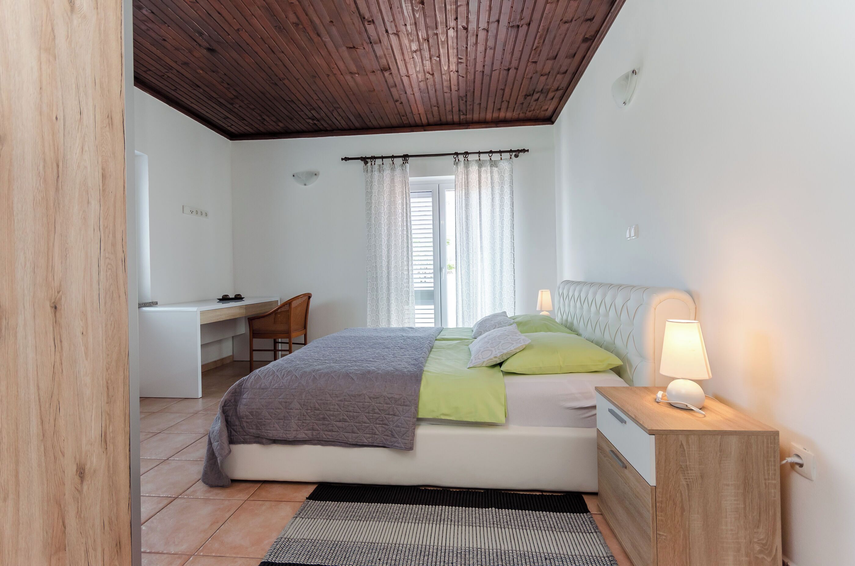 Una camera da letto con un letto, comodino, lampada, scrivania, sedia e una finestra con tende.