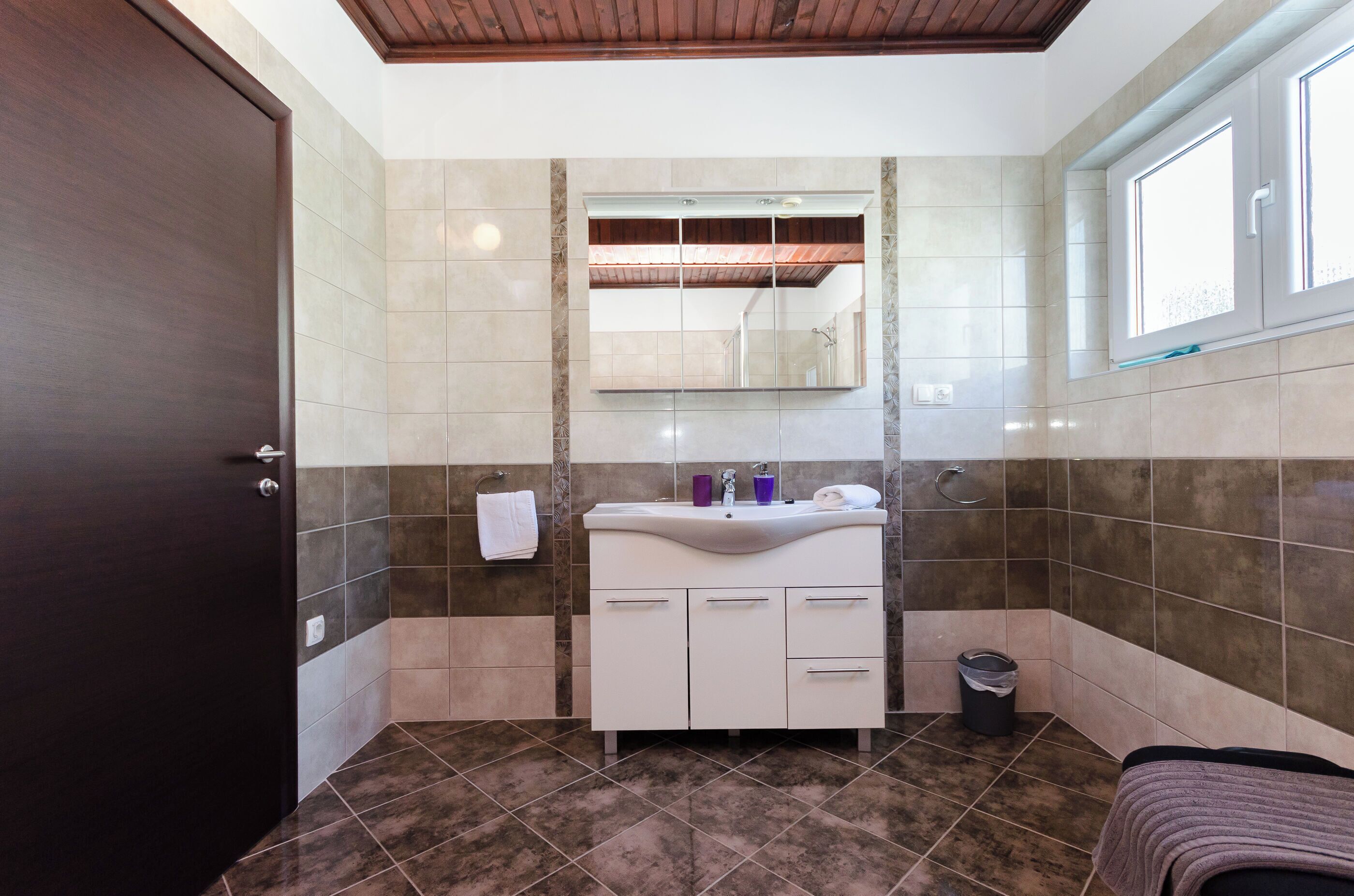 A bathroom with a dark wooden door, a white vanity with a mirror, and a window with a view of the outside.