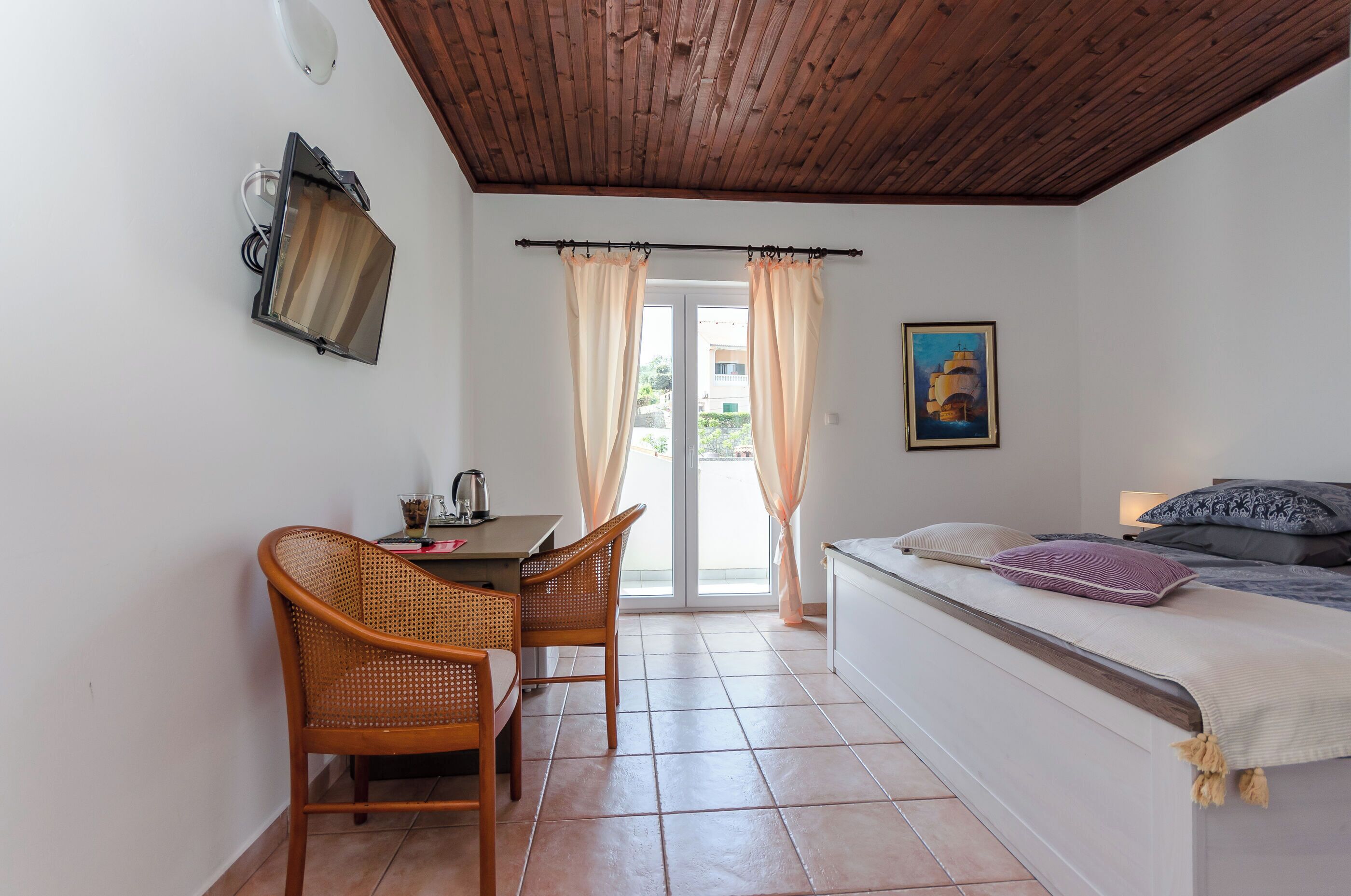 A room with a bed, a desk with chairs, a window with curtains, and a painting on the wall.