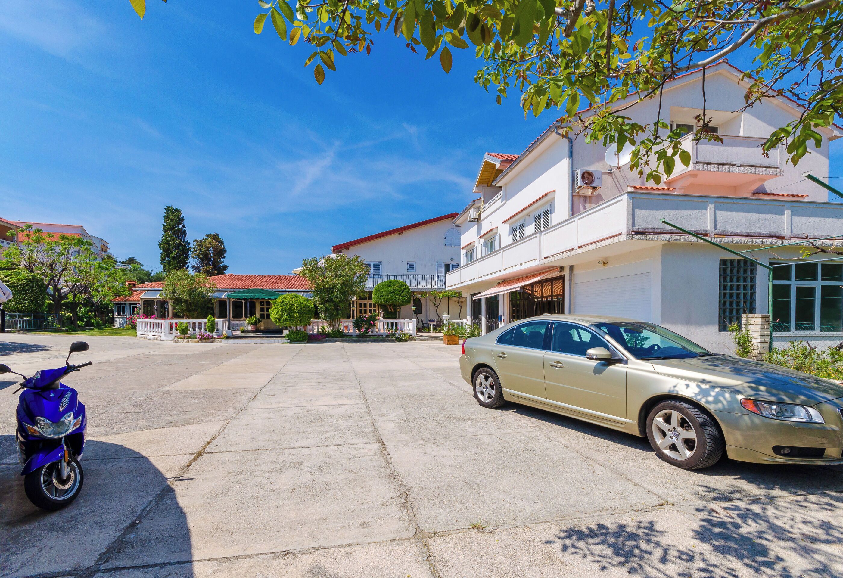 A residential area with a parked car and a scooter, houses with balconies, and a clear blue sky.